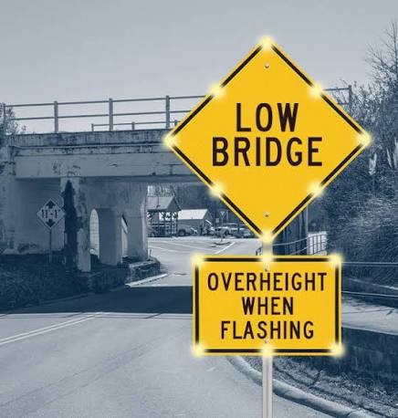 Illuminated LOW BRIDGE warning sign reading OVERHEIGHT WHEN FLASHING, activated by a Kestrel OHVD sensor near a low-clearance bridge